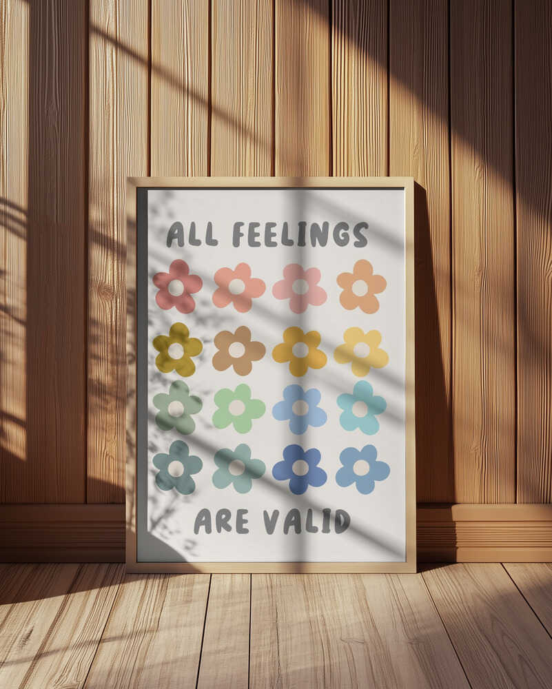 Framed pastel flower art print leaning against a wood-paneled wall with natural sunlight and shadows. Cute multicolor floral design with positive quote “All feelings are valid.” Calming and affirming nursery and kids room wall decor.