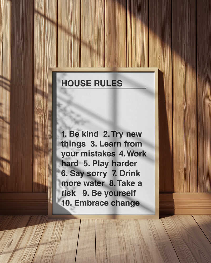 Modern typography poster titled “House Rules” featuring a motivational list with phrases like “Be kind” and “Embrace change.” Displayed in a light wooden frame leaning against a wooden wall, with natural sunlight and shadows. Minimalist black and white wall art for contemporary interiors.