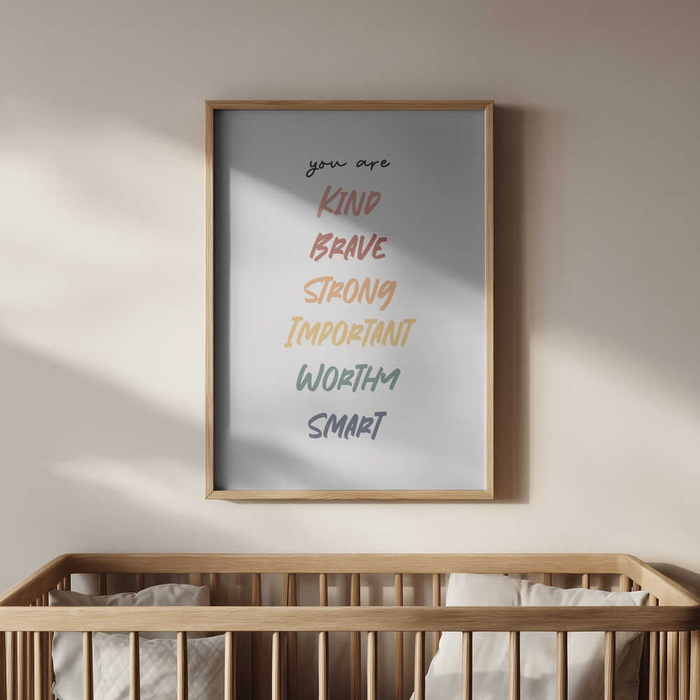 Framed “You are kind, brave, strong, important, worthy, smart” print hanging above a wooden crib. Gentle motivational typography creating a warm, supportive, and inspiring nursery environment.